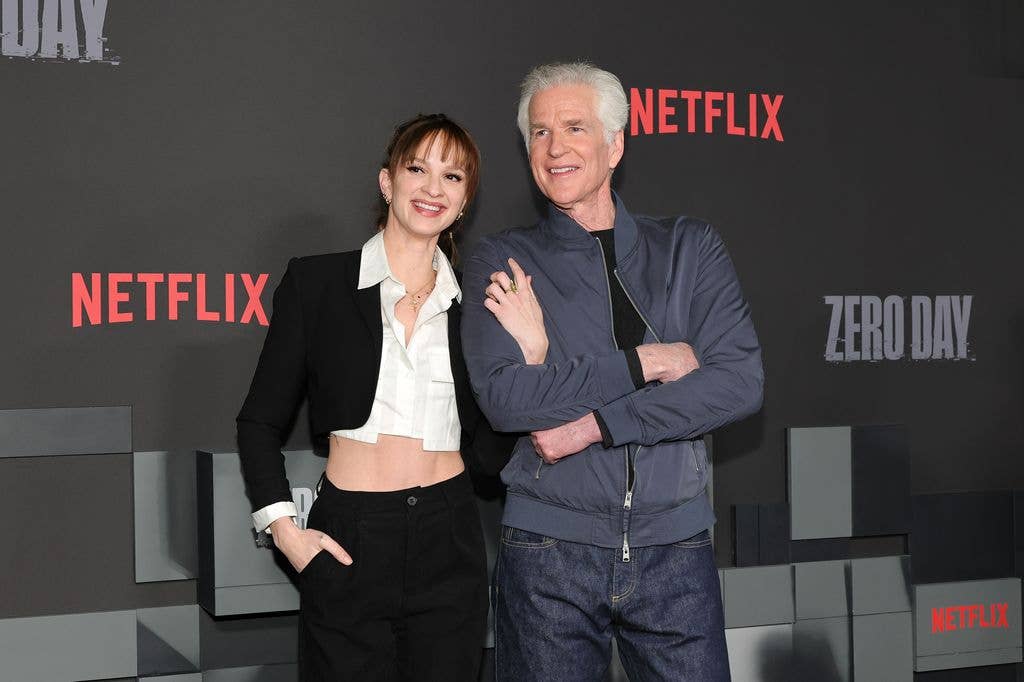 Ruby Modine with her father Matthew Modine. Ruby wears a cropped black jacket and crobbed black shirt with black jeans. She puts her right hand in her pocket and her left hooked up and through her father's crossed arms. Matthew wears grey jeans, a grey jacket, zipped at the bottom and a black tshirt.