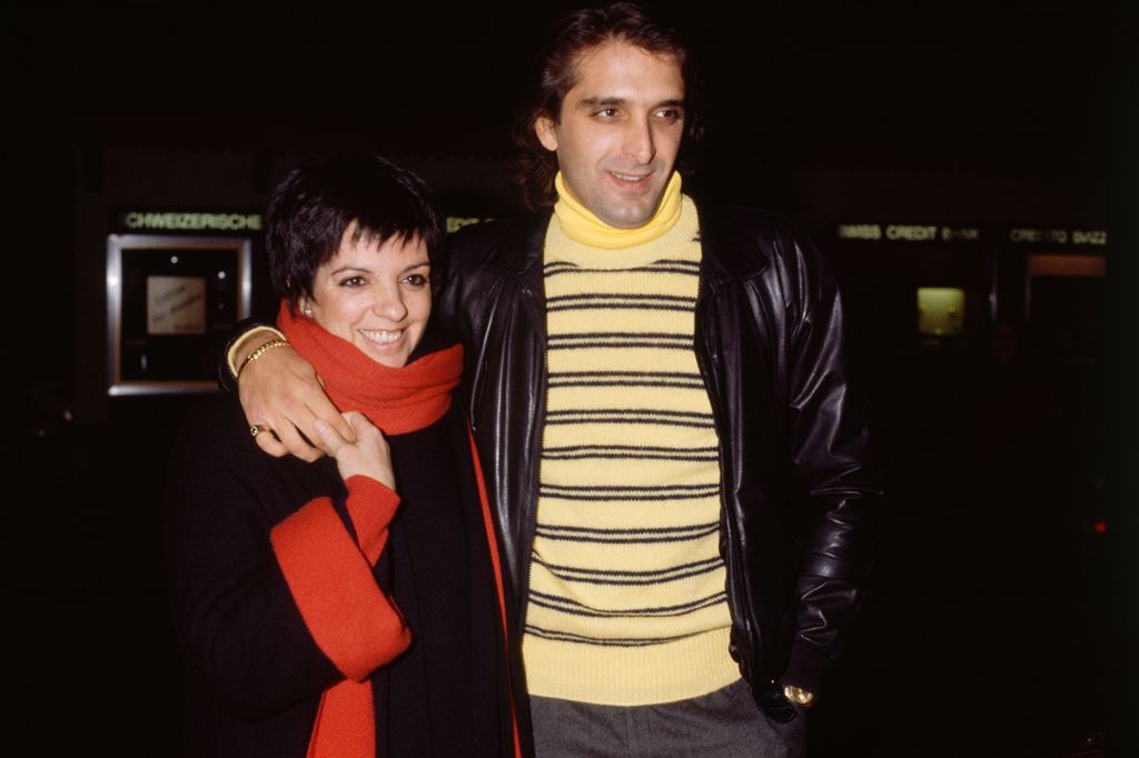 Liza Minnelli wears a black coat with red cuffs and holds onto her husband Mark Gero's hand which is wrapped around her shoulders.