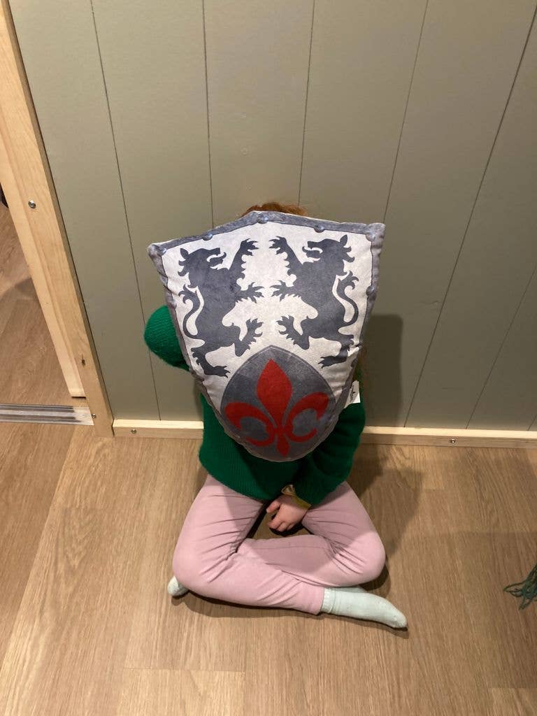 Small child sitting cross legged on the floor holding a shield-shaped cushion in front of her face