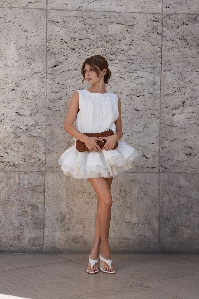 Olivia Jade wears brown hair in a low loose updo with a center part and face-framing tendrils, gold hoop earrings, gold rings, a brown suede clutch bag, a white sleeveless dress with a pleated front bodice and a voluminous multi-layered white lace and tulle mini skirt, white leather thong sandals shoes with mid-height block heels, outside AJE, during Paris Fashion Week - Womenswear Spring Summer 2026