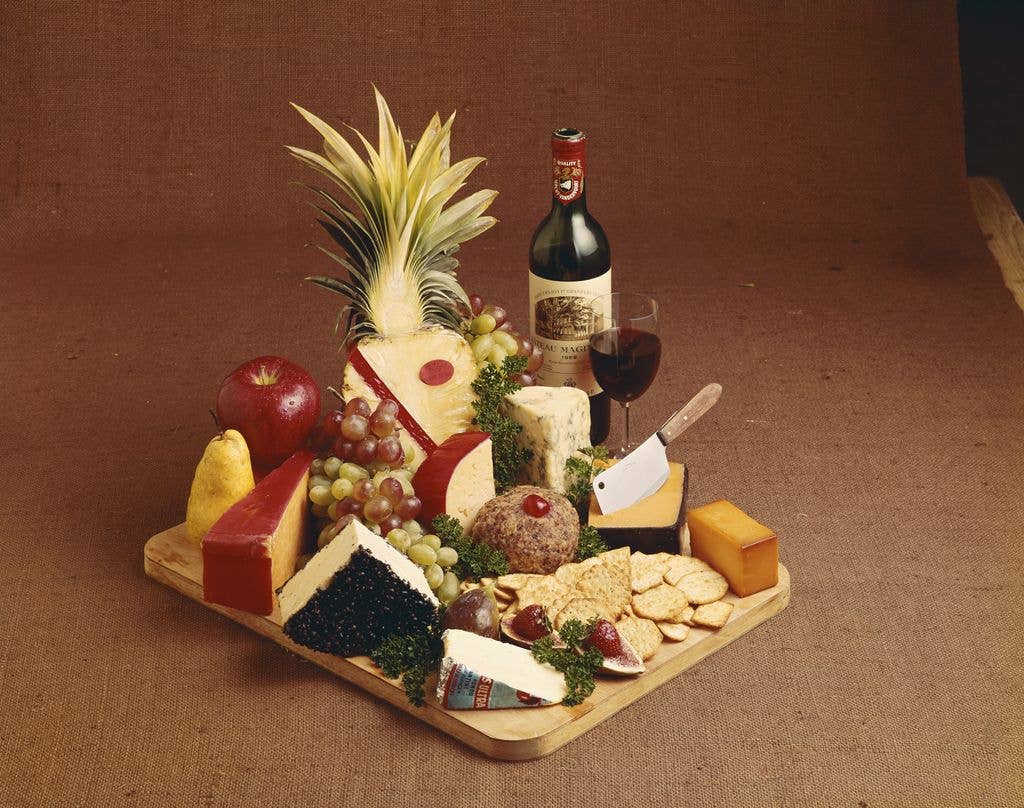 Various cheese, fruits and wine on chopping board with a plain brown backdrop.