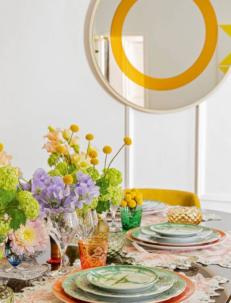 The kitchen table is set with colourful flowers in the centre and patterned green plates, complemented by a clear mirror with an orange circle on the wall