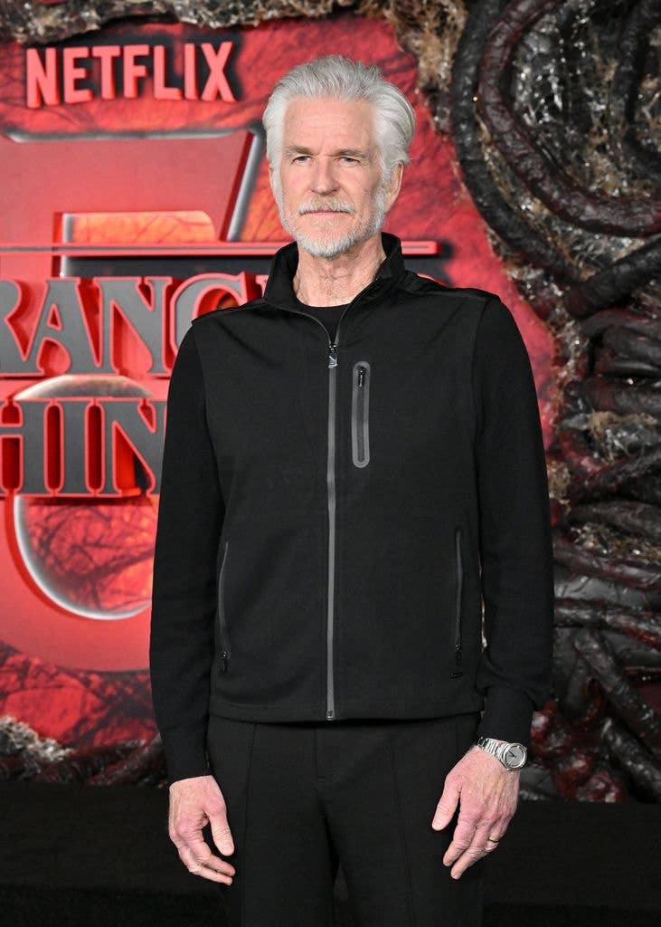 Matthew Modine in a black zipped jacket standing in front of a promotional backdrop for Netflix's Stranger Things season five.