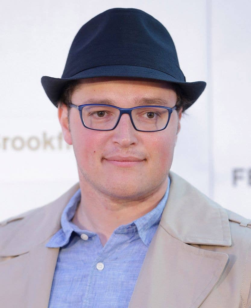 Boman Modine in a black fedora, glasses, a blue shirt and a beige jacket.