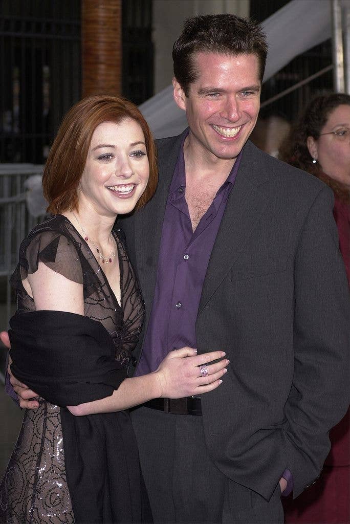 Alyson Hanigan wears a black dress with a black shawl around her elbows and holds a hand to Alexis' stomach. Alexis wears a grey suit with a purple shirt open at the neck. Both look to the right of the image.