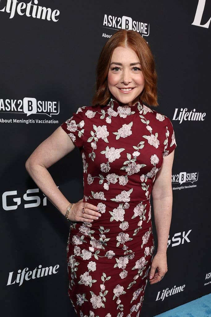 Alyson Hannigan stands with one arm by her side and the other hand on her hip, wearing a high necked red dress with a bold white floral pattern. Her hair is cut to her shoulders and she smiles at the camera.