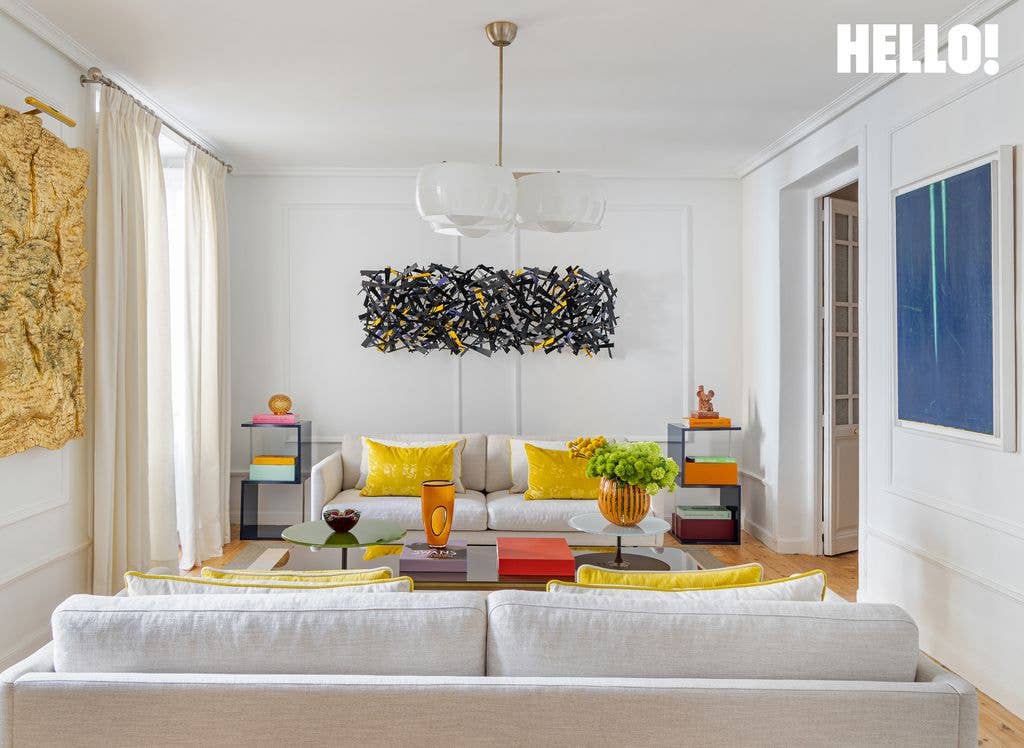 A white-walled living area in designer Silvia Tcherassi’s Madrid home features art pieces, white sofas with yellow cushions, and a white chandelier