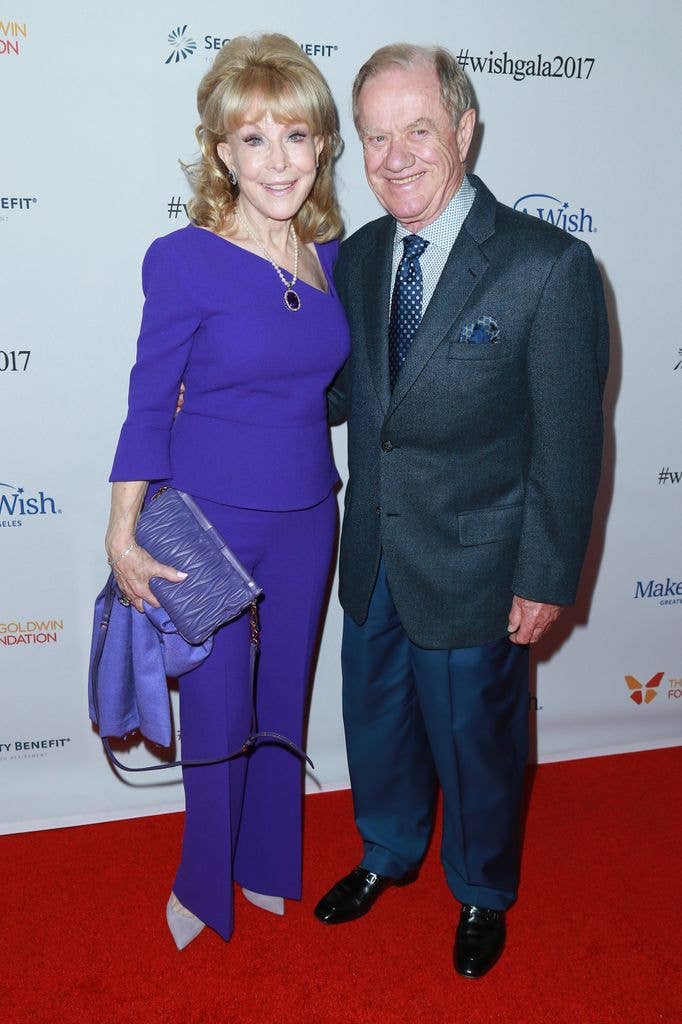 Barbara Eden wears a purple top and purple trousers with grey pointed heels, standing next to husband Jon Eicholtz who wears a dark textured suit jacket with navy trousers and a navy pocket square. 