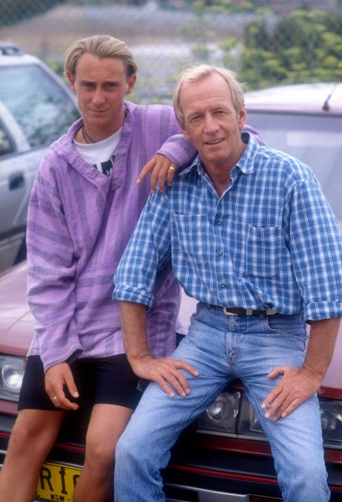 Paul Hogan sits on the bonnet of a car in jeans and a check shirt, holding his hands on his legs. His son Scott sits next to him in shorts and a purple shirt, resting his left elbow on his father's shoulder.