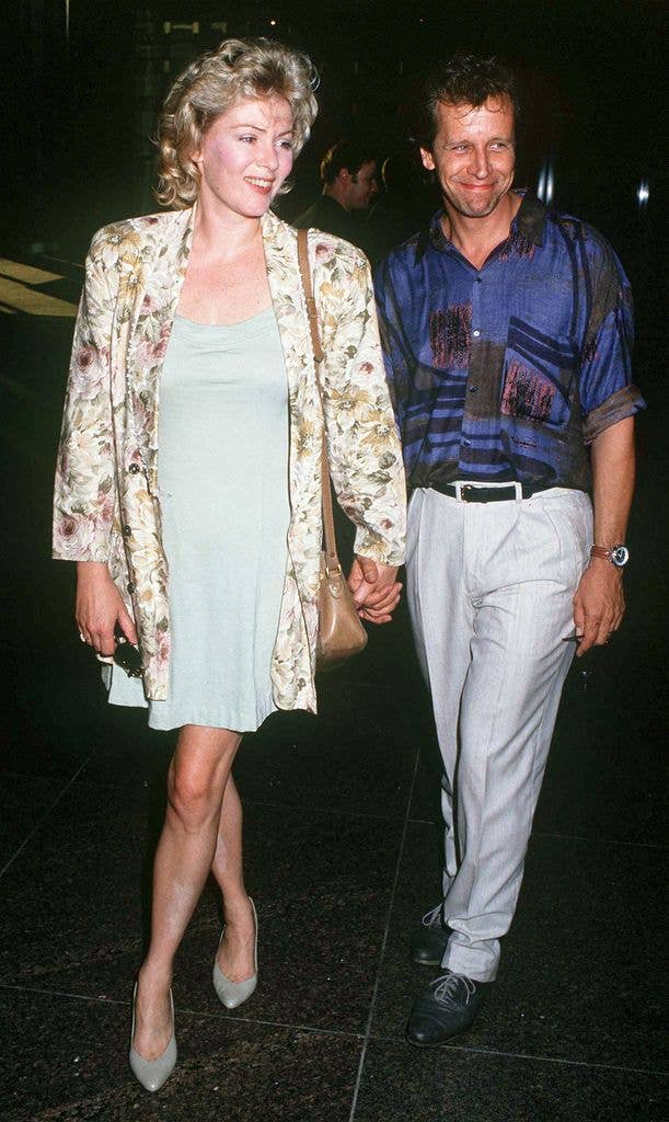 Jean Smart wears a mini light blue dress with a floral jacket, a brown shoulder bag and pointed shoes. She holds husband Richard Gilliland's hand, Richard wears a purple patterned shirt with grey trousers and black dress shoes.