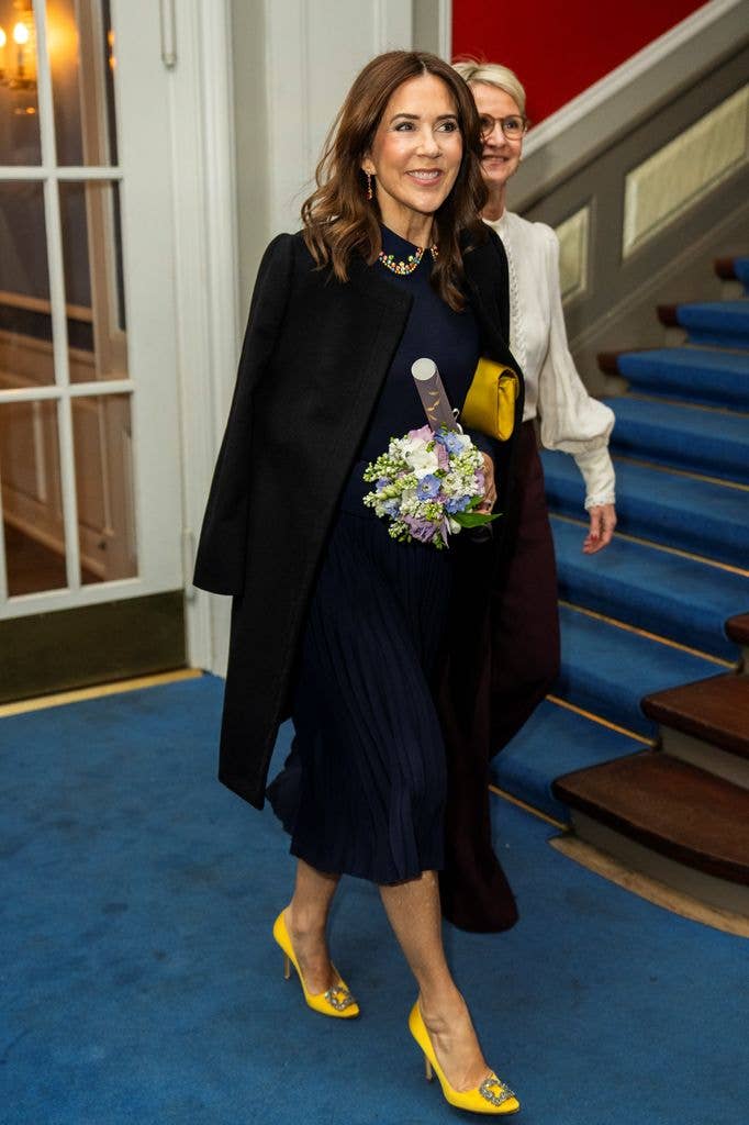 Queen Mary in navy dress and yellow heels holding flowers 