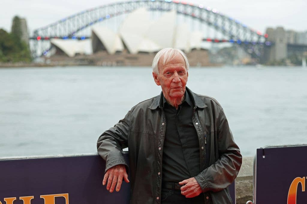 Paul Hogan leans back against a banner for Crocodile Dundee: The Encore Cut, resting his elbow on top of the barrier. 