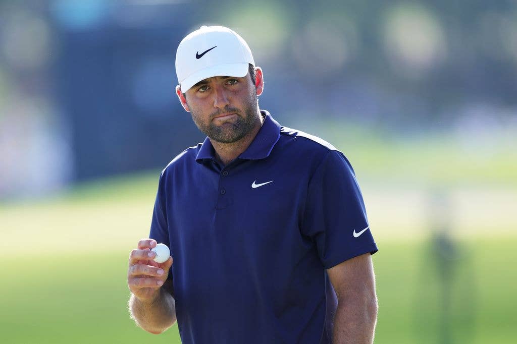 Scottie Scheffler wears a navy polo with a white Nike logo and a white hat with a navy Nike logo. He holds up a golf ball in his right hand, his right arm bent at the elbow.