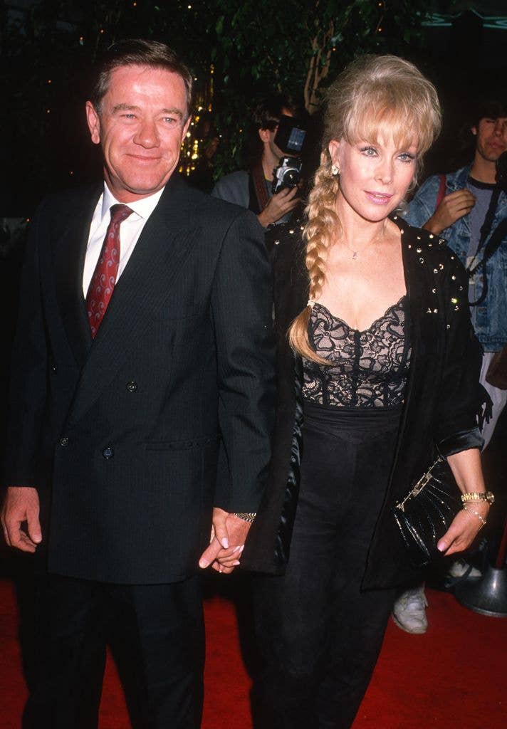Jon Eicholtz holds hands with wife Barbara Eden,. He wears a dark suit with a red tie while she wears a dress with a structured, nude lined bodice and black cardigan, holding a clutch against her side.