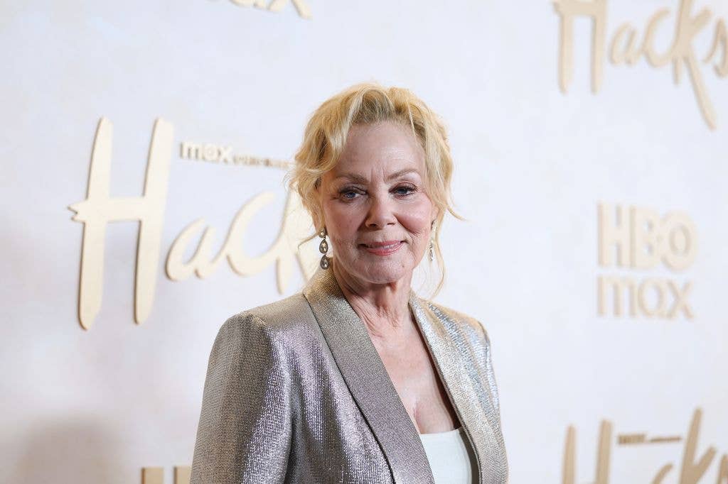 Jean Smart wears a silver jacket over a white top in front of a promotional backdrop for HBO Max series 'Hacks'.
