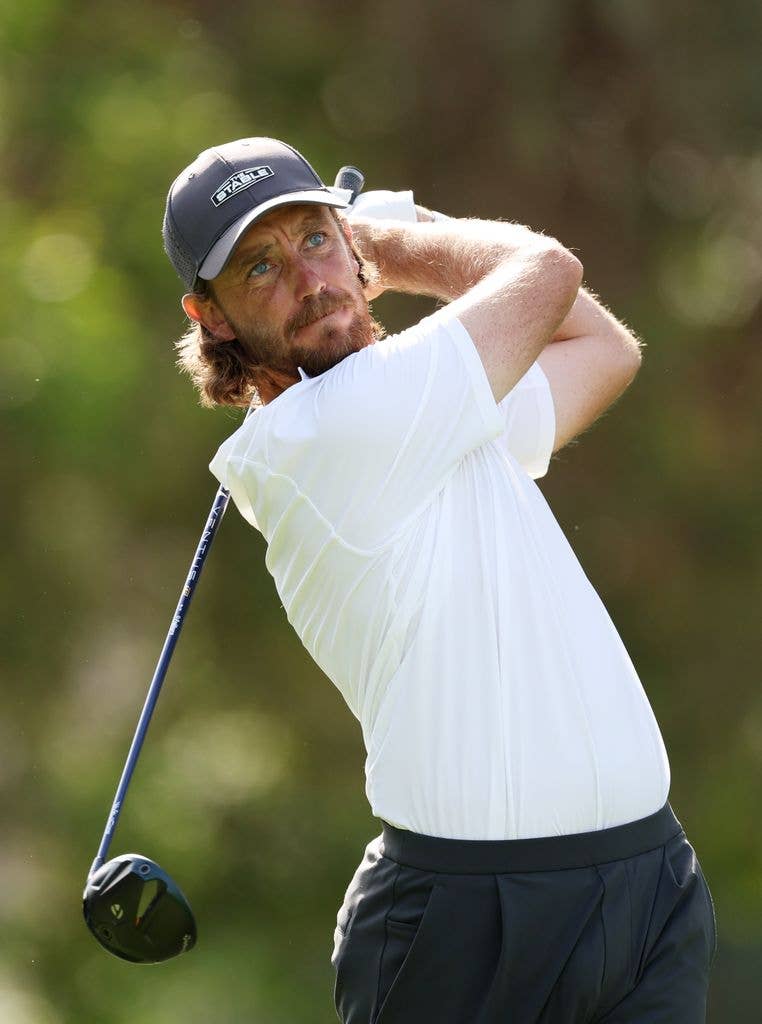 Tommy Fleetwood holds a golf club in two hands over his shoulder and behind his back. He wears a grey cap, white short sleeved shirt and grey trousers. 