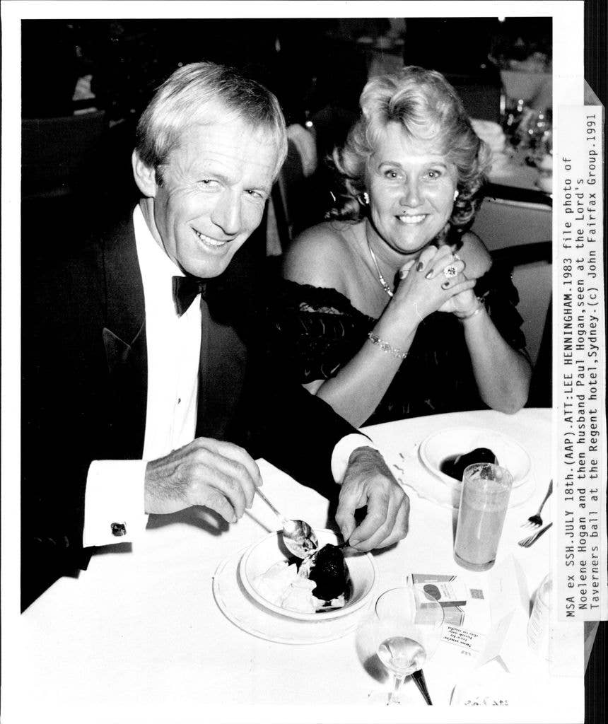 Noelene Hogan and then husband Paul Hogan, seen at the Lord's Taverners ball in formal wear. Paul wears a suit and bowtie while eating a dish of dessert while Noelene smiles at the camera with her clasped hangs held under her chin.