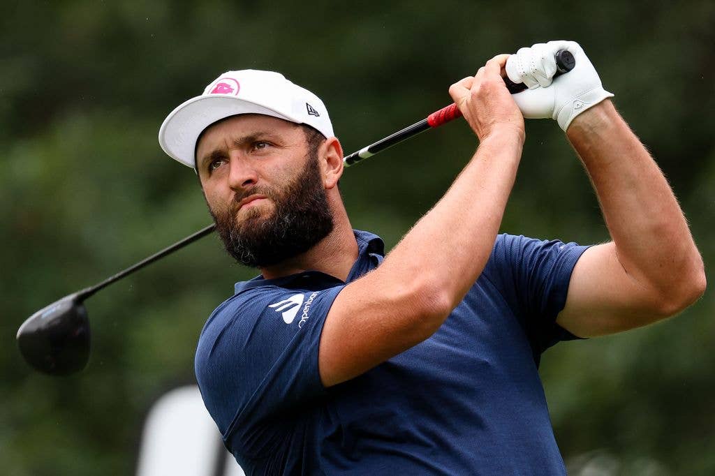 John Rahm looks to the top left of the image and holds his golf club over his left shoulder and back behind his neck. He wears a white cap, blue polo shirt and a single white glove on his left hand.