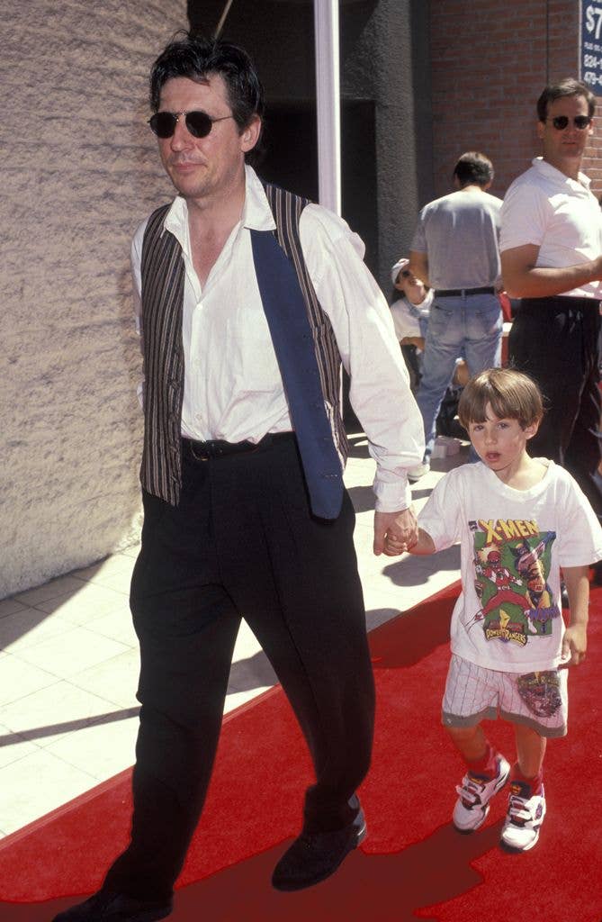 Gabriel Byrne wears opaque sunglasses, an open striped waistcoat with a grey lining, white shirt tucked into dark trousers and walks along the red carpet, holding son Jack's hand. Jack wears an X men tshirt, shorts and white trainers with red socks.