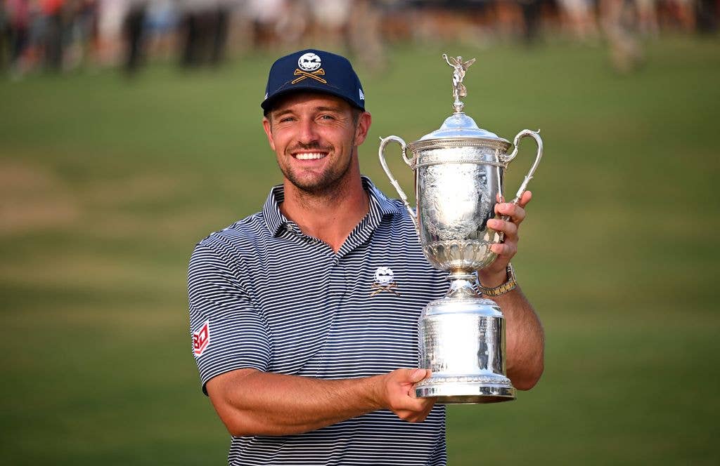 Bryson DeChambeau holds the silver US Open trophy in his left hand and supports the base with his right. He smiles in a navy striped pop and a navy hat, both emblazoned with a skull and crossbones logo.