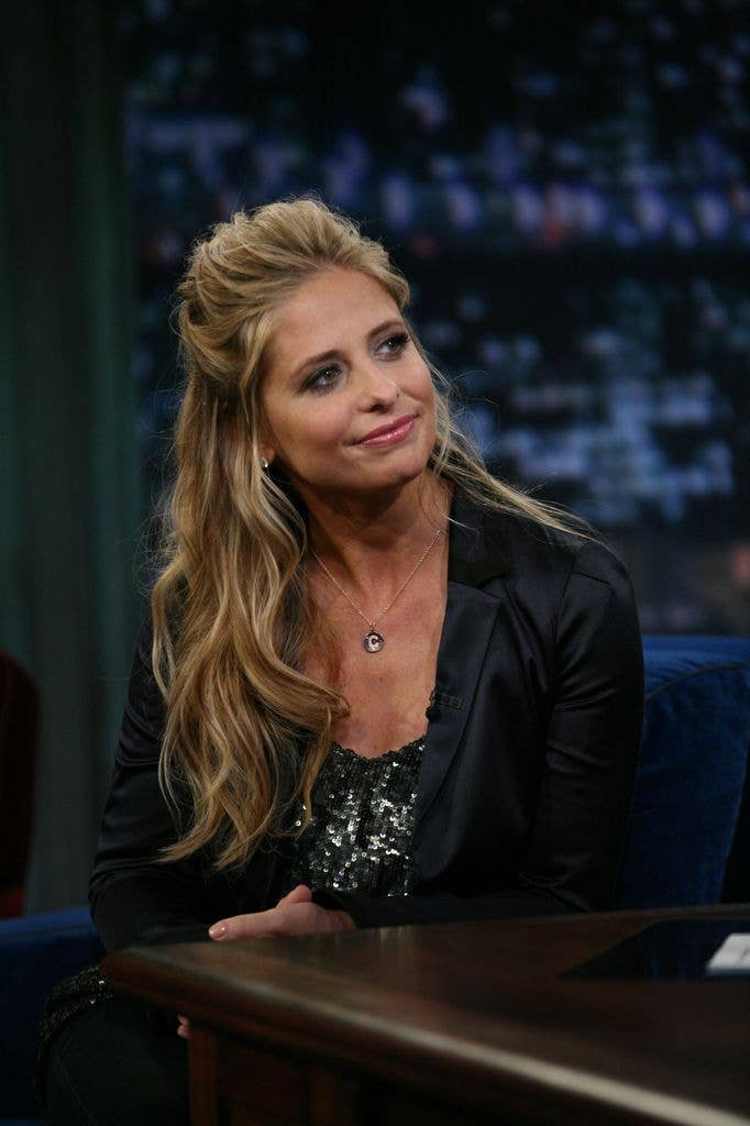 Sarah Michelle Gellar sat next to a desk in a sparkly black dress with a black jacket, tilting her head to the left of the image with some of her blonde curly hair pulled up and to the back of her head.