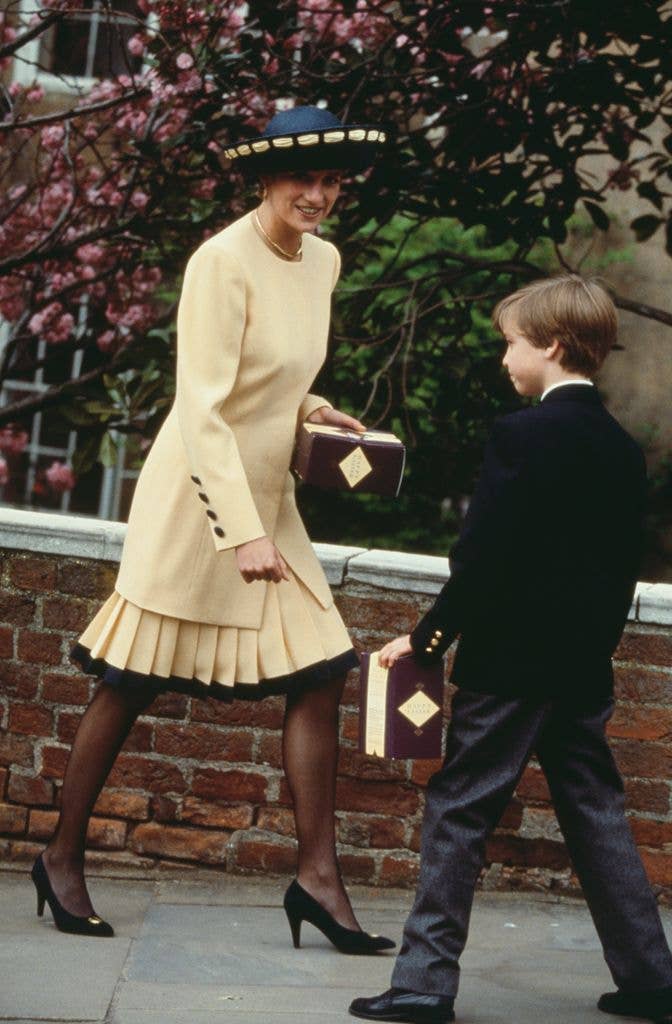 Princess Diana smiles at her son Prince William while wearing a butter yellow pleated suit with black trim, black pumps, sheer tights, and a wide-brimmed navy hat adorned with yellow ribbon.
