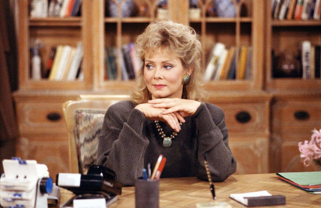 Jean Smart in a dark brown jumper sits in a chair and rests her elbows on a wooden desk, folding her hands and looking to the left of the image.