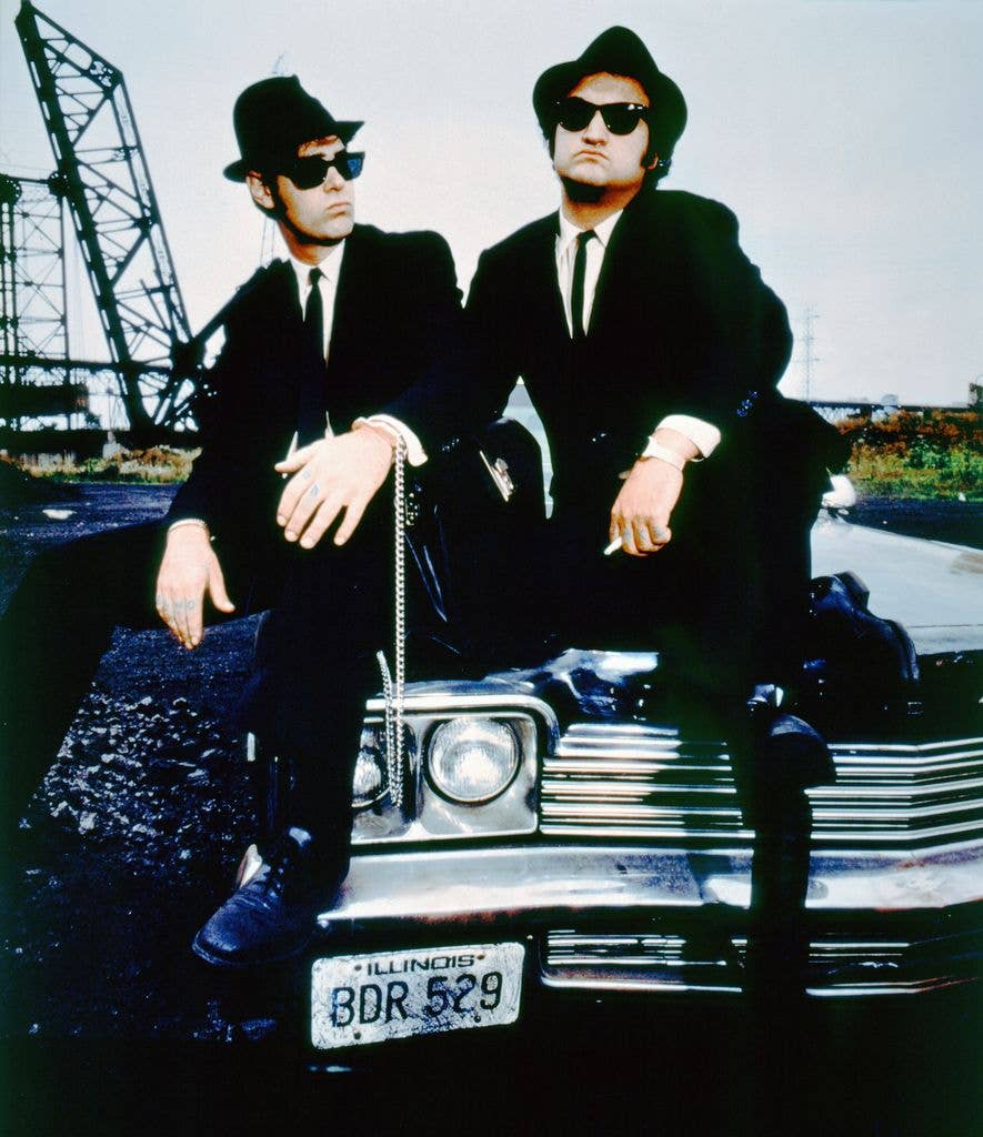 Dan Aykroyd and John Belushi wear hats, sunglasses and suits and sit on the bonnet of a car.