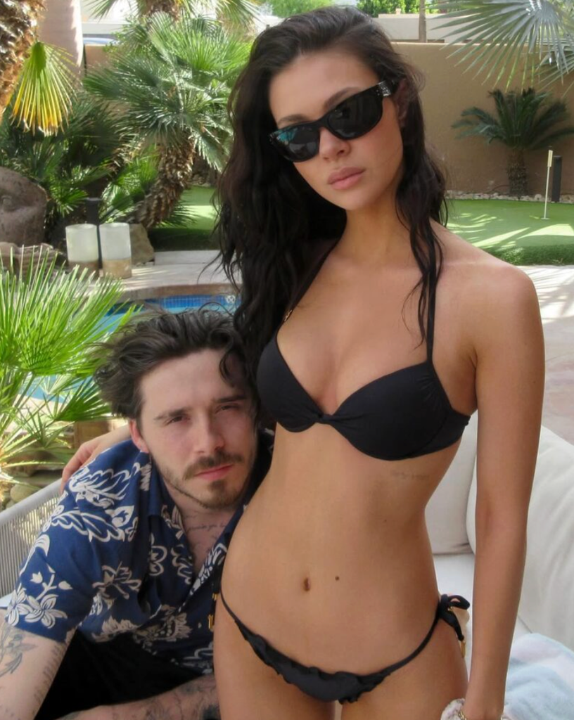 Nicola Peltz posing wearing a two-piece black bikini and shades with Brooklyn Beckham sat next to her with his arms around her