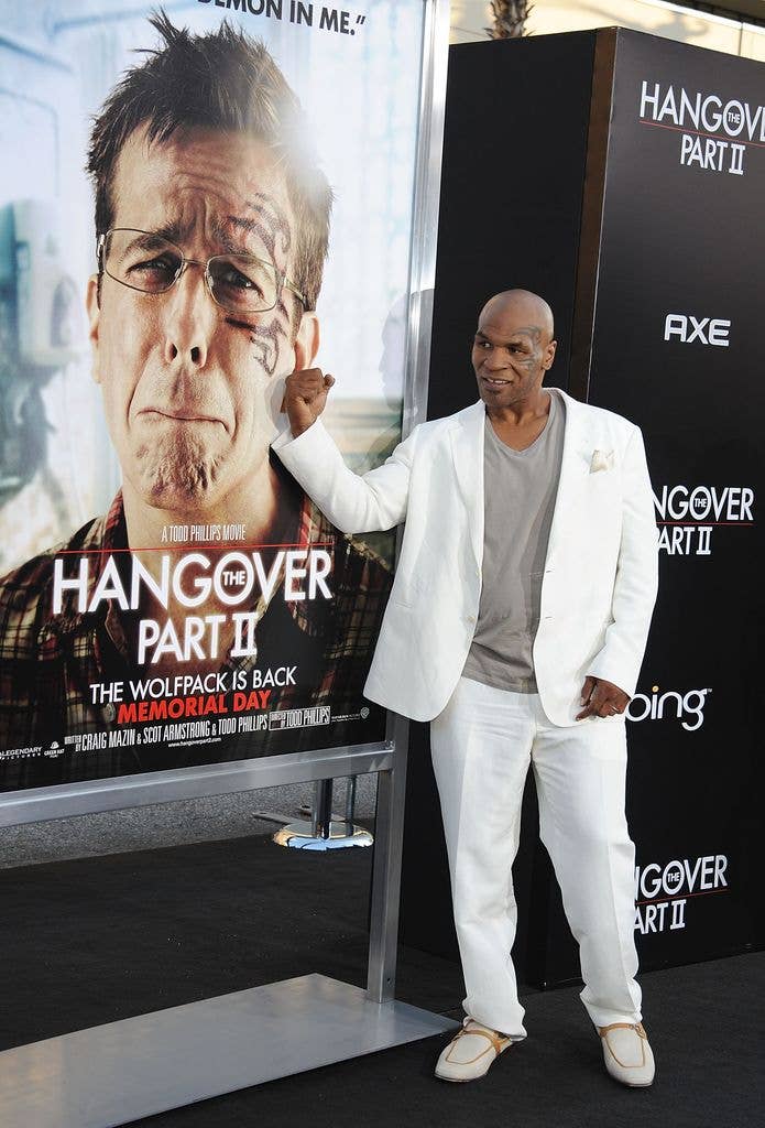 Mike Tyson wears a white suit and a grey tshirt, holding a fist up to a poster of Stu from 'The Hangover' who has the same tattoo on his face as Tyson.
