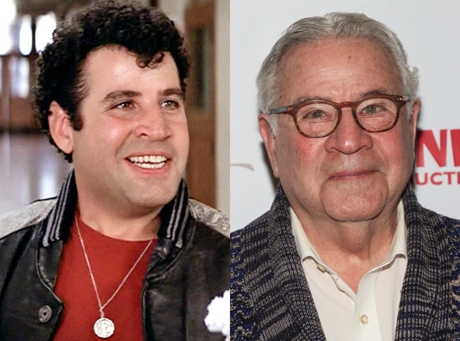 Split image of Michael Tucci. On the left, speaking to a teacher off screen, wearing a red top, silver penny necklace and black leather jacket with a quiff and curly black hair, looking to the right of the image. On the right, a modern photo of the actor wearing a striped knit cardigan and a cream shirt with brown glasses and short grey hair looking straight at the camera with a slight smile.