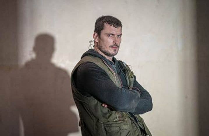 Person in green vest and black shirt stands with arms crossed, looking serious.