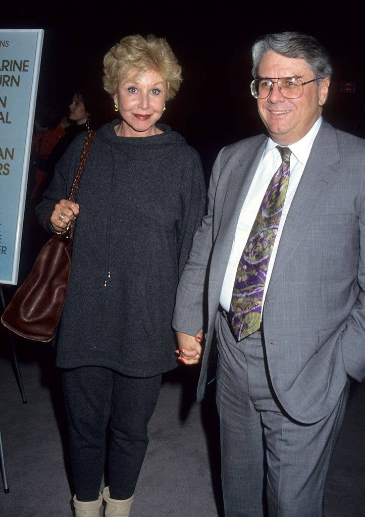 Michael Learned in a loose grey top with dark trousers and a brown shoulder bag holding hands with husband John Doherty who wears a grey suit and large glasses.