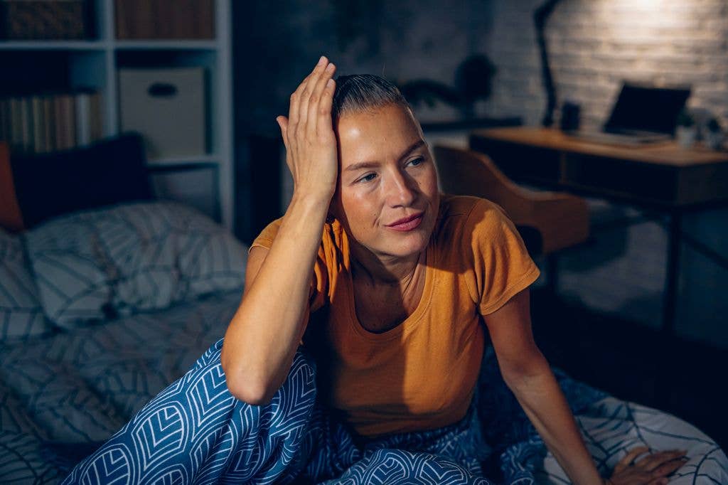 A woman with her hair pulled back and wearing an orange t-shirt sits on her bed at night. She has one hand on her head, looking away from the camera in a dimly lit bedroom, representing middle-of-the-night insomnia