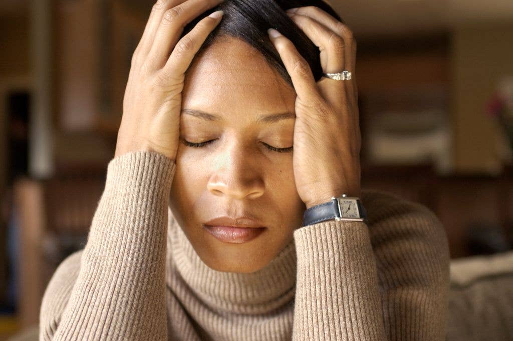 A woman in a beige turtleneck holds her head, eyes closed, wearing a watch and ring.