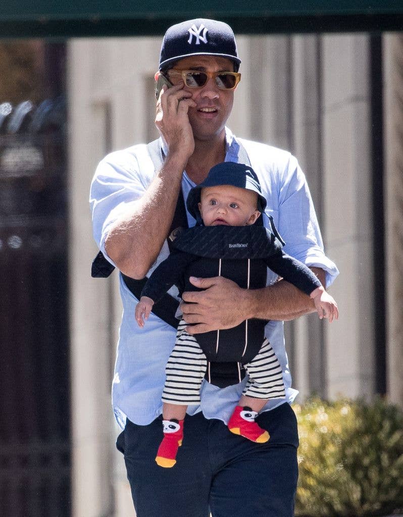 Bobby Cannavale holds his phone to his ear and holds his other arm around son Rocco who is strapped into a baby sling around his front.