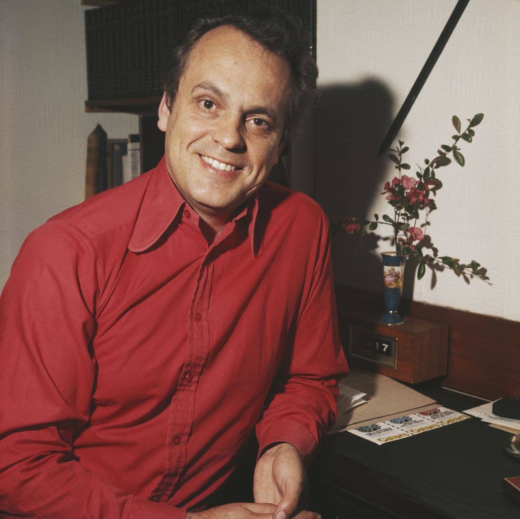 A smiling man in a red shirt sits at a desk with a vase of flowers.