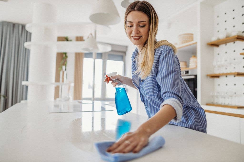 A woman in a blue shirt is cleaning a kitchen counter with a blue cloth and spray bottle.
