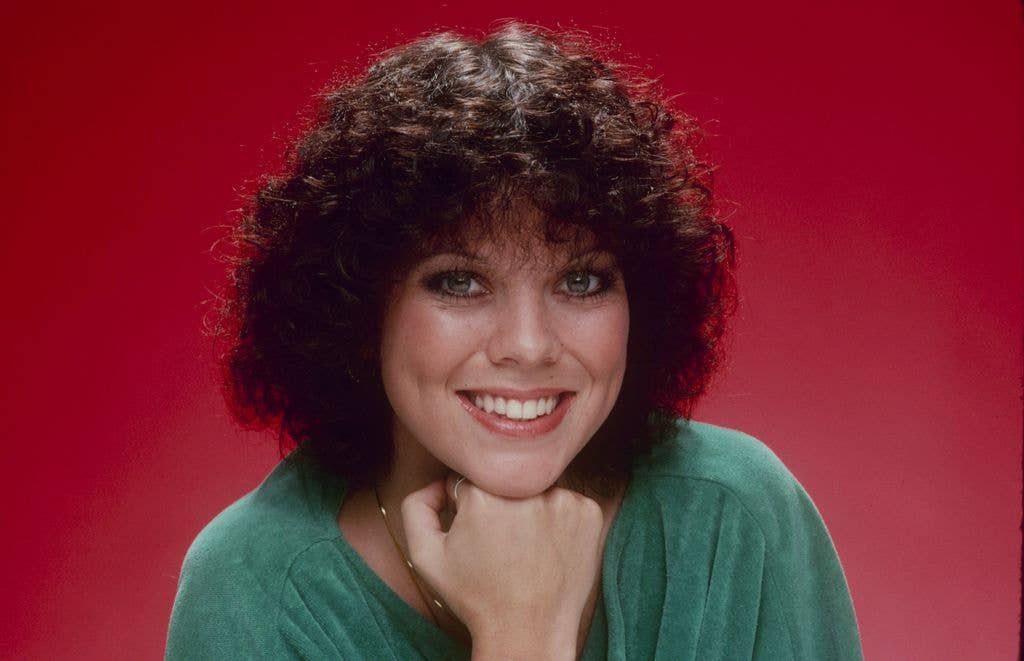 Erin Moran in a promotional headshot for Happy Days wearing a green top and resting her head on her hand against a red backdrop.
