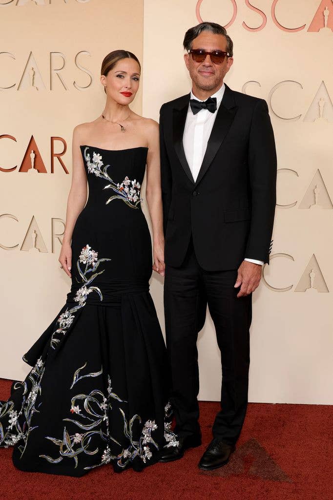 Rose Byrne and Bobby Cannavale at the Oscars in 2026. Rose wears a strapless black dress with a layered mermaid tail style and floral print. Bobby wears a tux and bowtie. 