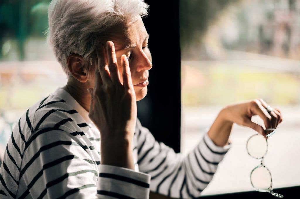 A person with short white hair, wearing a black and white striped shirt, holds glasses and touches their face.