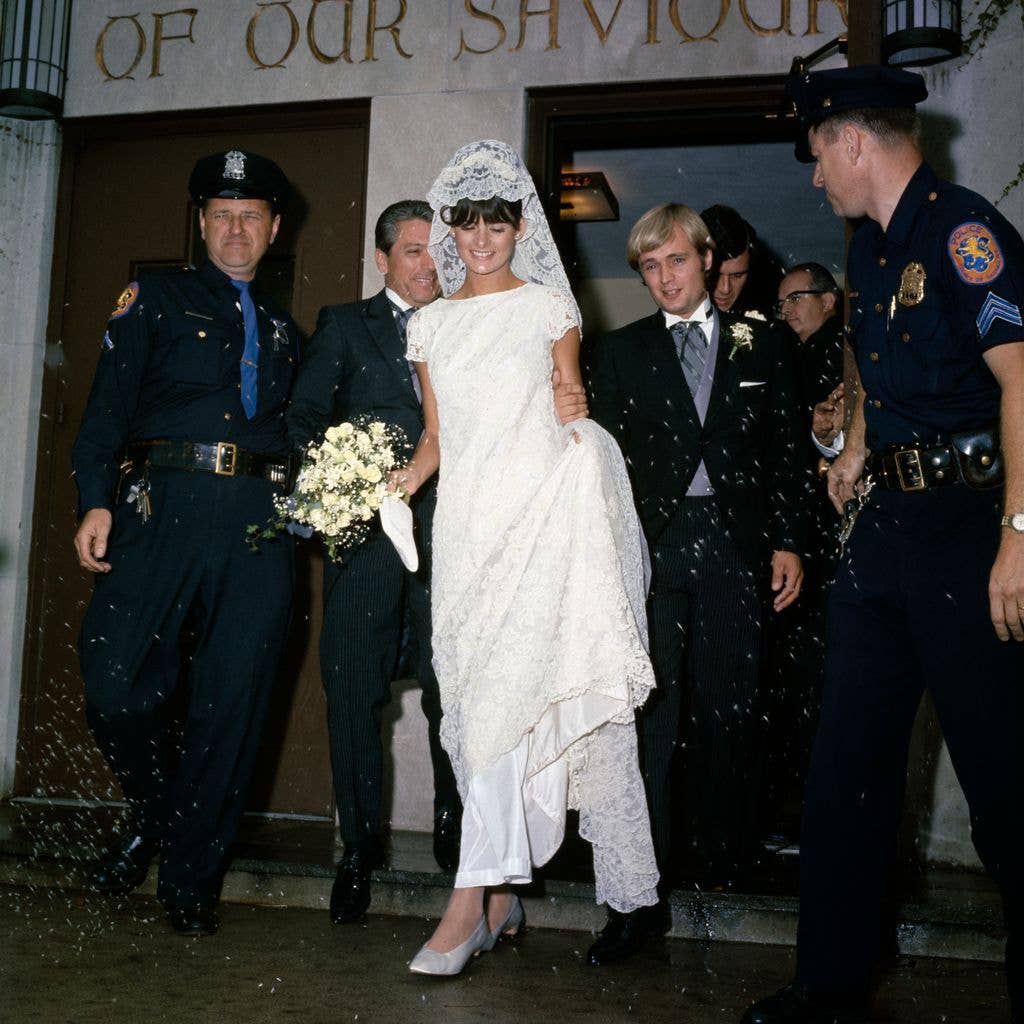 Katherine Carpenter wears a wedding dress with short sleeves and a high neckline, veil and holds a bouquet of white roses. She leaves the Lutheran Church of Our Saviour with husband David McCallum escorted by police. 