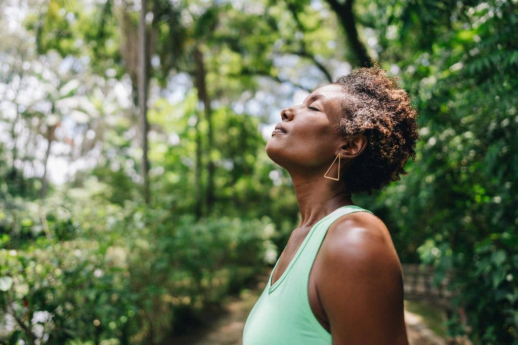 A woman in a green tank top looks up, eyes closed, in a forest setting.