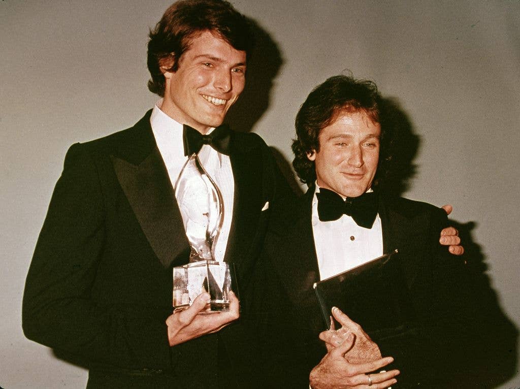 American actors Christopher Reeve and Robin Williams pose backstage at the People's Choice Awards in tuxes. Christopher holds an award and wraps his arm around Robin's shoulders, while Robin holds a black folder.