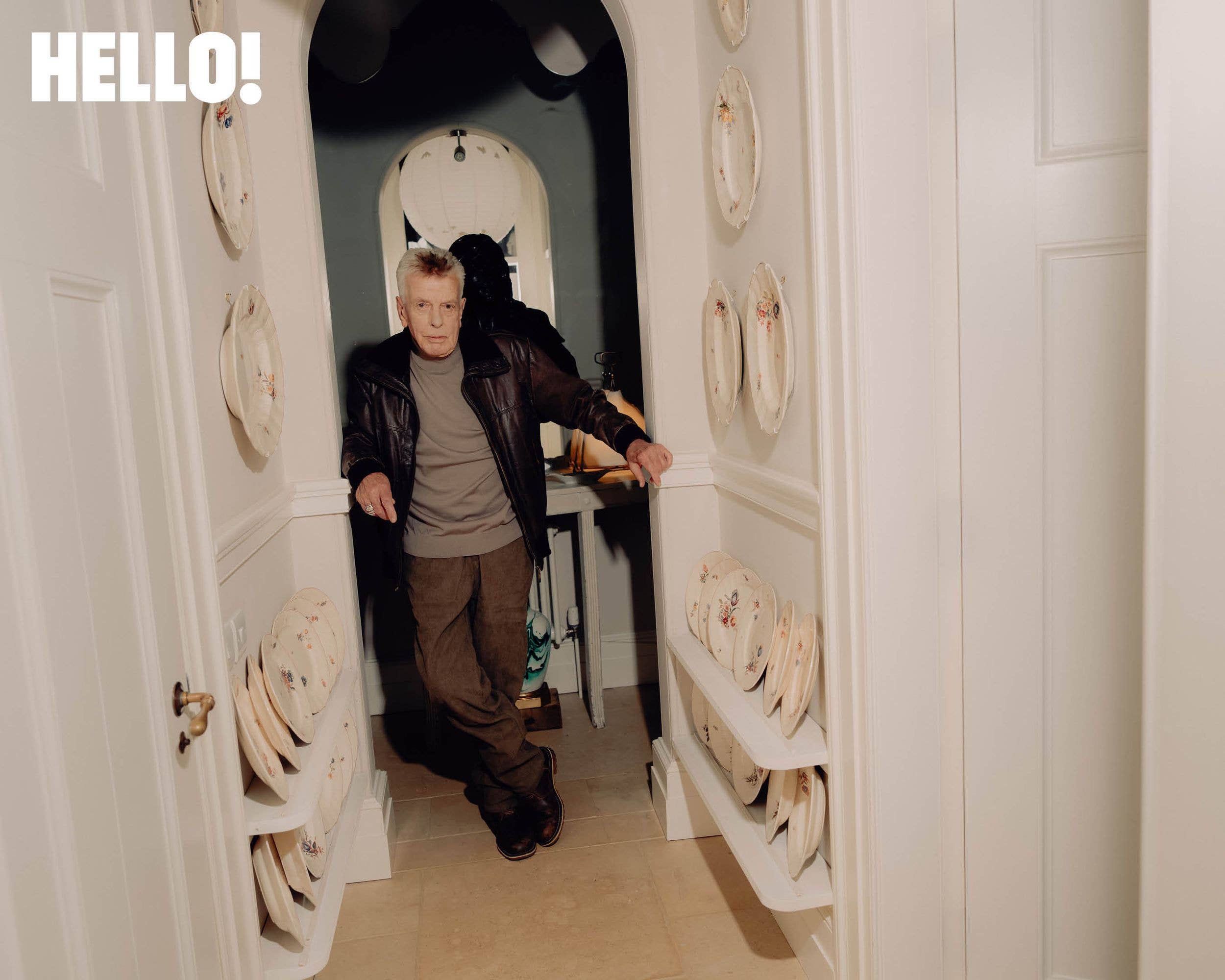 Nicky Haslam leans against the archway in the hallway of his home in the Cotswolds surrounded by ornamental plates
