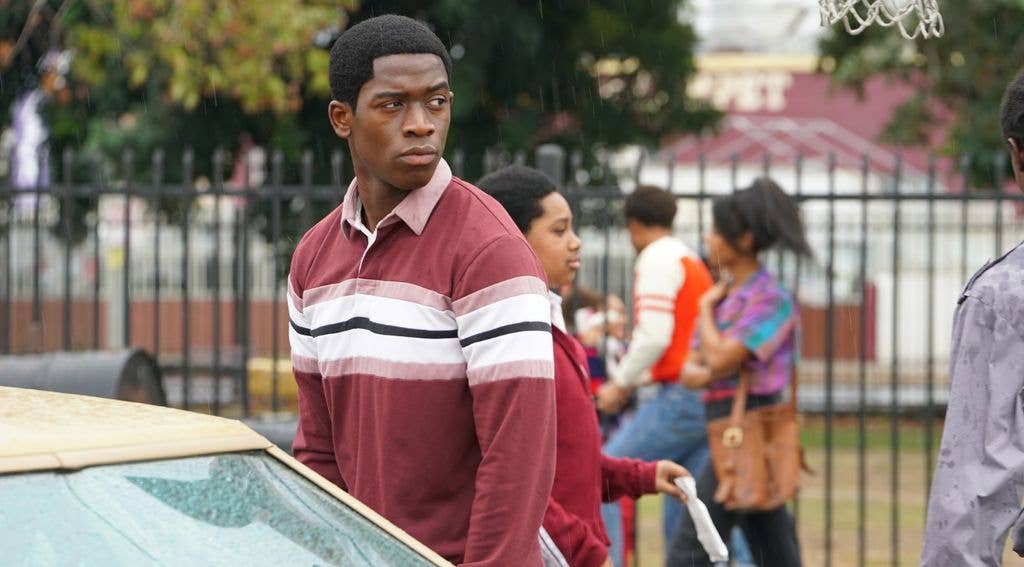 Damson Idris stands in the rain near a car, wearing a maroon and white striped shirt.