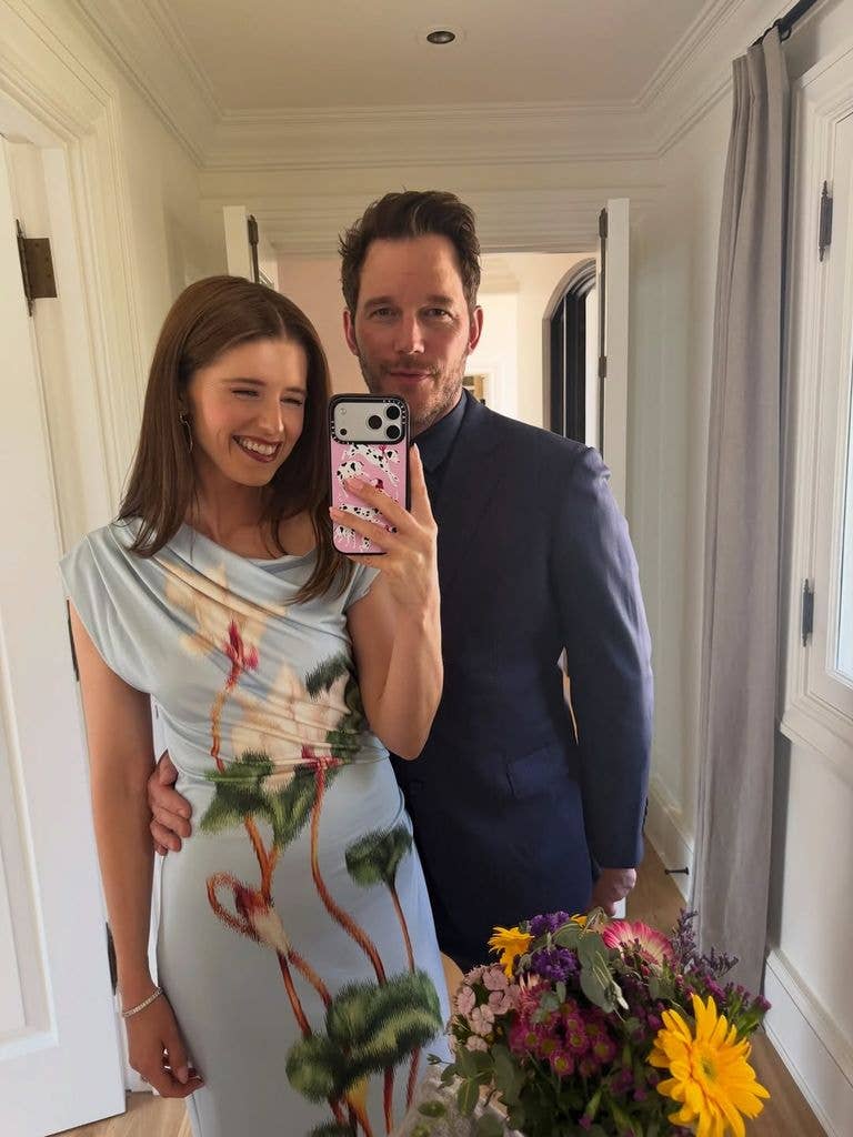 A mirror selfie of Katherine Schwarzenegger and Chris Pratt standing together indoors. Katherine is smiling and holding a smartphone with a pink patterned case to take the picture. She is wearing a light blue sleeveless top with a large floral pattern. Chris stands close behind her with an arm around her waist, wearing a dark blue suit jacket and a dark dress shirt.