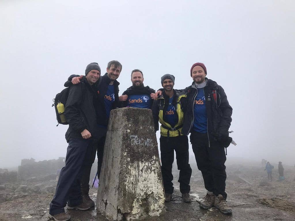 Hikers round a trig point on a grey and mistyu mount,ain