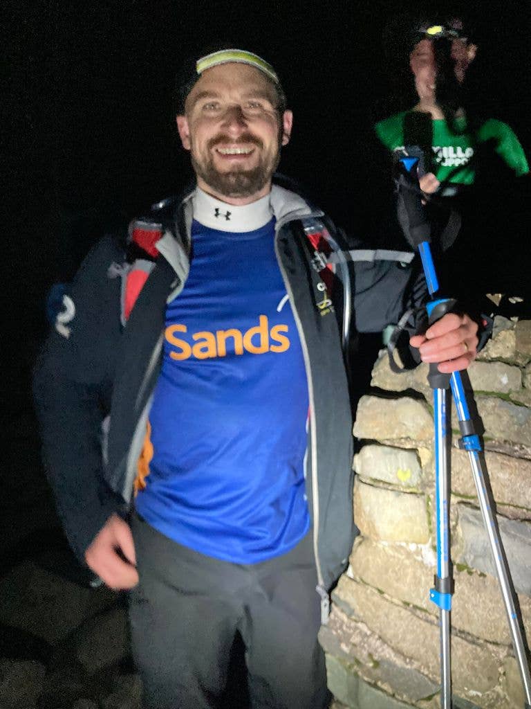Man in hiking gear wearing blue Sands t-shirt and holding poles in the dark