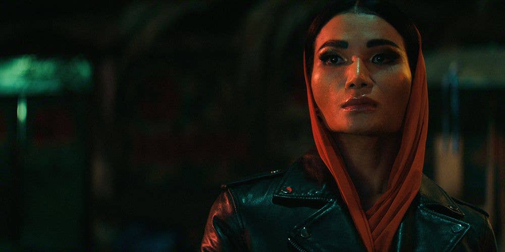 A woman in a black leather jacket and orange scarf stands in a dimly lit setting.